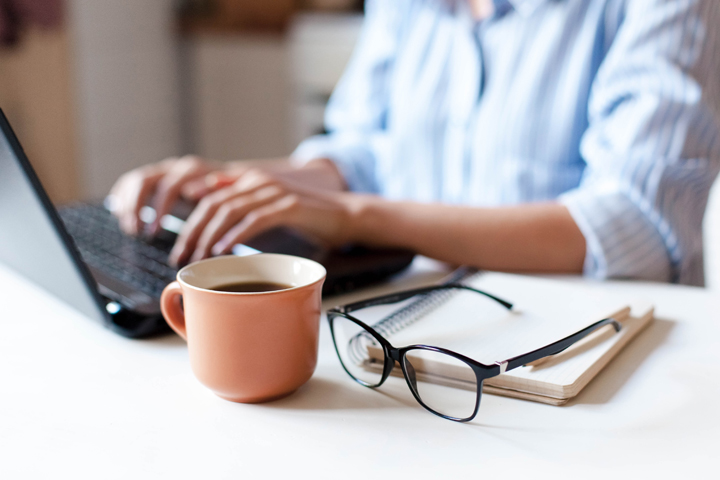 Photo of someone using a laptop with a hot drink and glasses on their desk